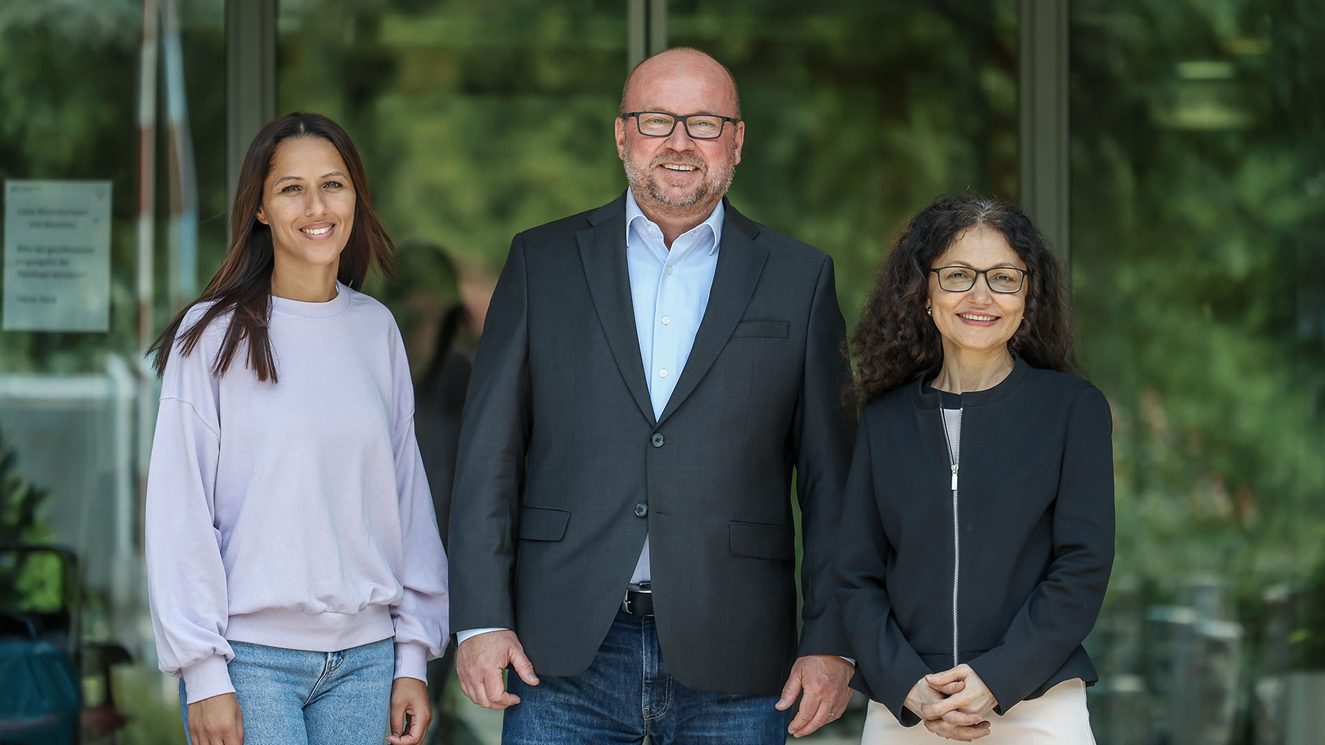 Chahrazad Hsini, Dieter Ritter und Sevgi Eker stehen vor dem Eingang des Gemeindestifts. Neben dem Eingang stehen hohe grüne Büsche und Bäume. 