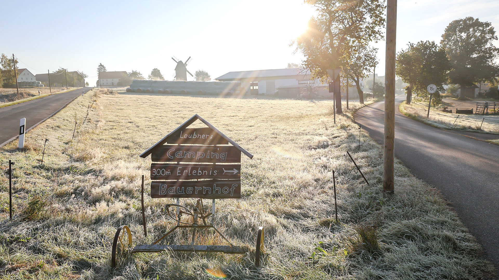 Wintersonne fällt auf ein mit Raureif bedecktes Feld. Im Hintergrund ist im hellen Dunst eine alte Windmühle zu sehen. Vorne steht ein Hinweisschild auf einen Erlebnisbauernhof.