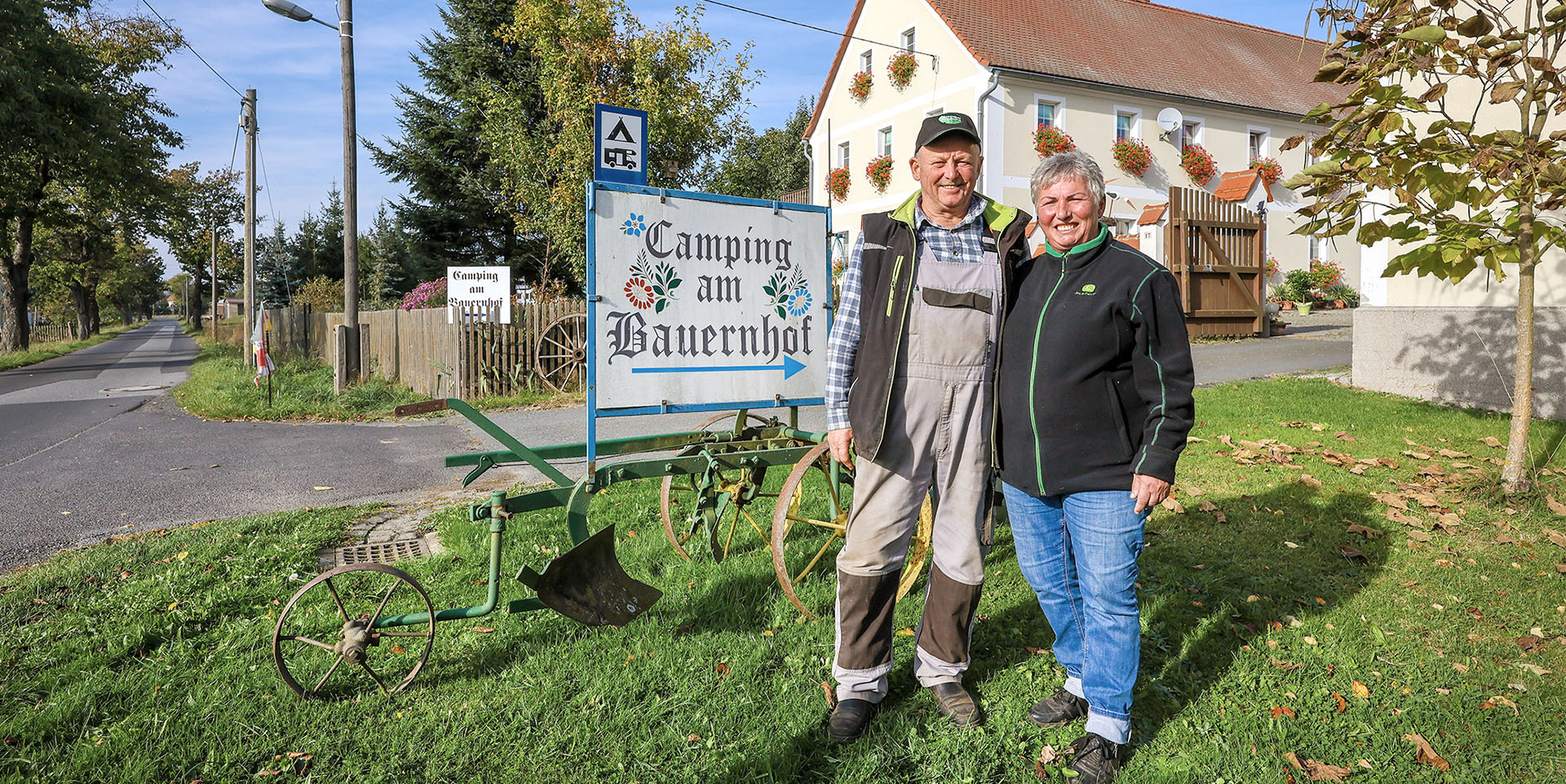 Die Leubners vor ihrem Bauernhof Ein älteres Ehepaar in Arbeitskleidung steht vor einem geraniengeschmückten Haus. Auf einem Schild hinter ihnen steht, „Ferien auf dem Bauernhof”..