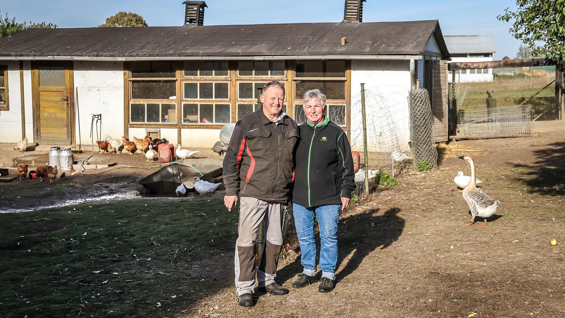 Ein älteres Ehepaar, der Mann links, die Frau rechts, stehen vor einem Geflügelstall mit Hühnern und Gänsen.
