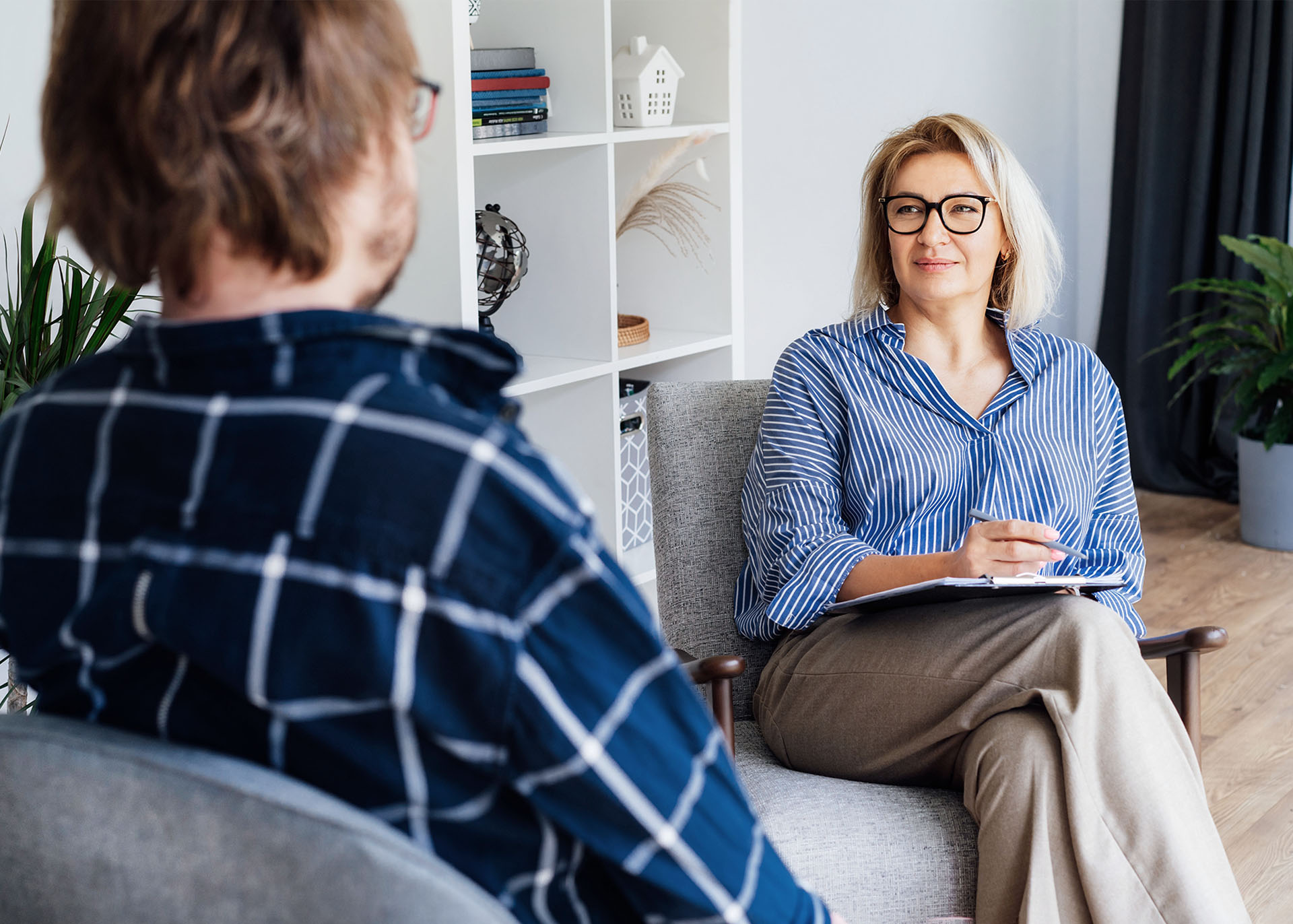 Eine Frau sitzt in einem Büroraum in einem Sessel, auf ihrem Schoß liegt ein Klemmbrett. Sie schaut ihren Gesprächspartner freundlich an. Der Gesprächspartner ist nur schräg von hinten am linken Bildrand zu sehen. Im Hintergrund steht ein offenes weißes Regal.