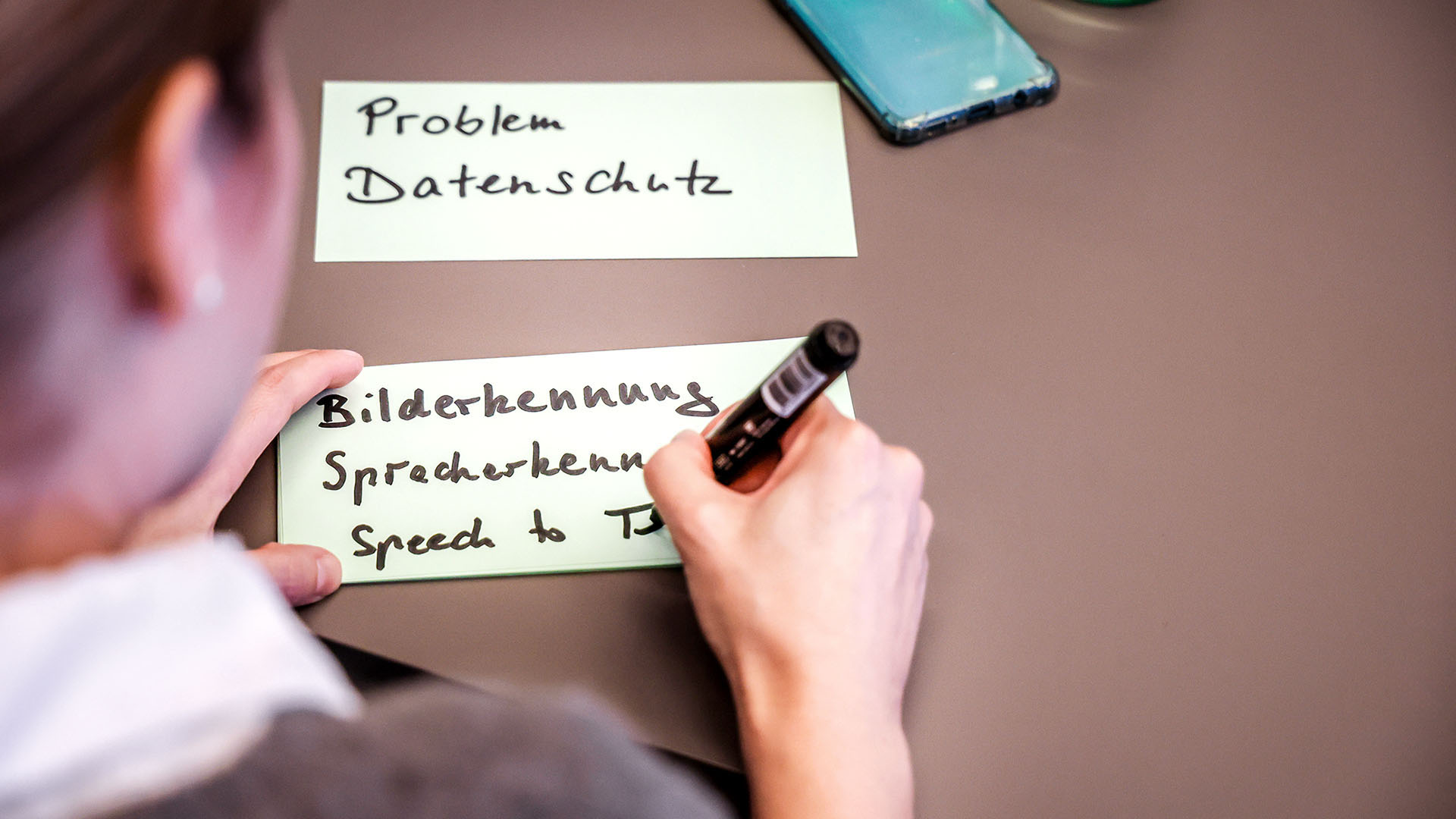 Eine Frau schreibt etwas auf eine gelbe Haftnotiz. Der Blick der Kamera fällt über ihre Schulter auf die Notiz.