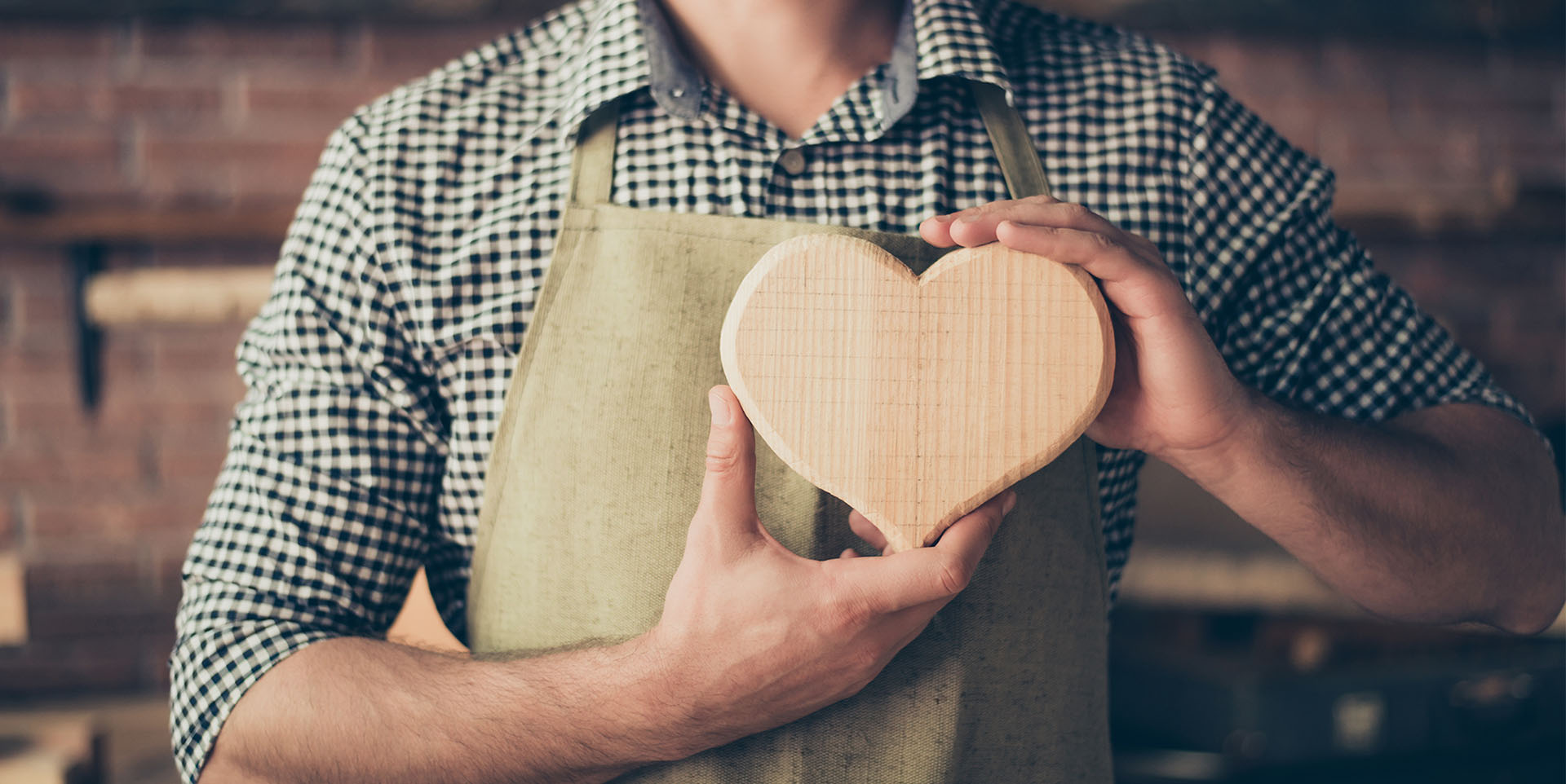 Jobcarving Zu sehen ist der Oberkörper eines Mannes in einem kariertem Hemd, darüber trägt er eine weiße Schürze. In seinen Händen auf Höhe des Herzens hält er ein Holzherz vor seine Brust.