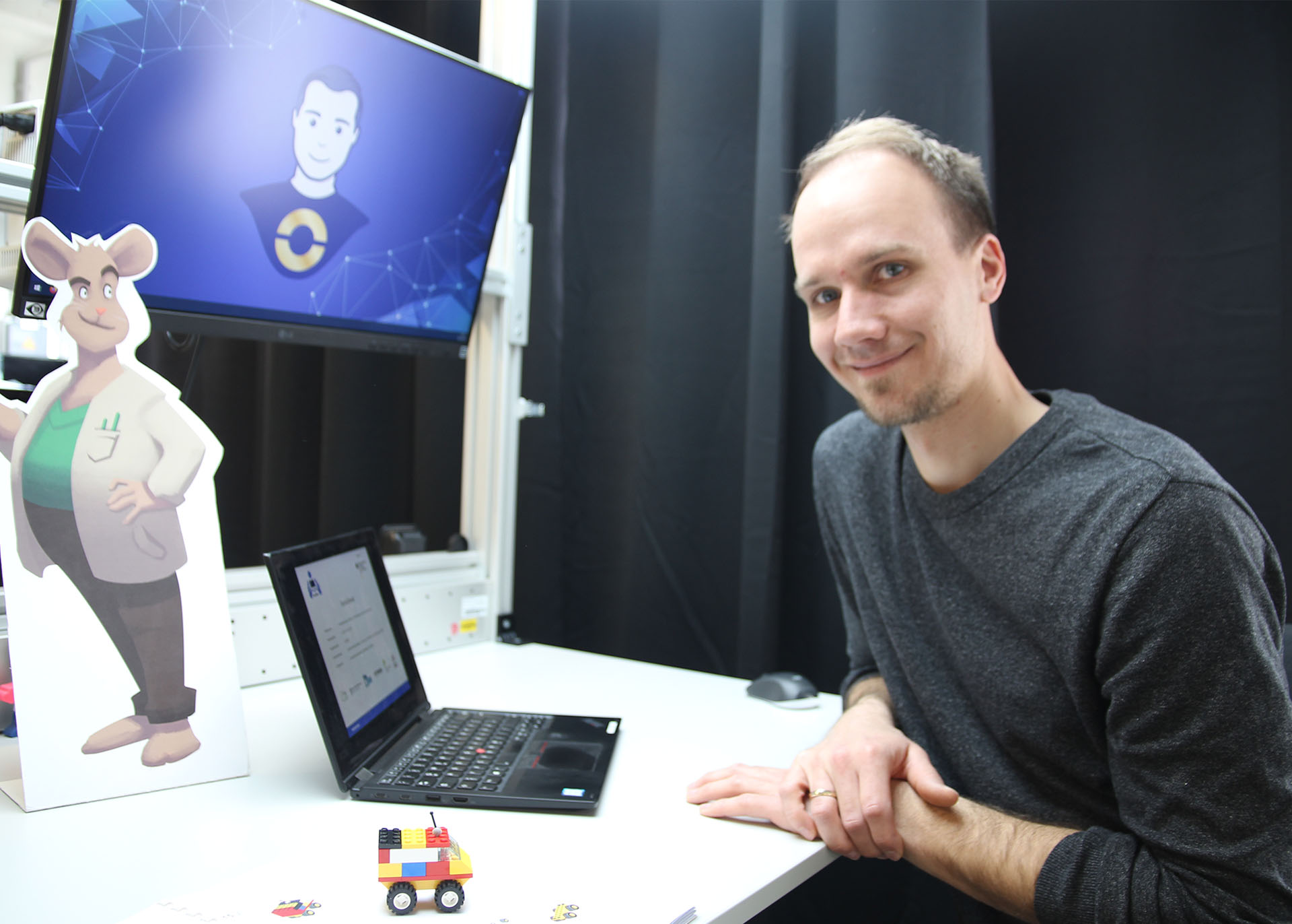 Der Projektkoordinator sitzt am Arbeitstisch. Darauf ein Laptop, ein Aufsteller der Comic-Maus Gregor im Anzug und das H-Auto aus Legosteinen.