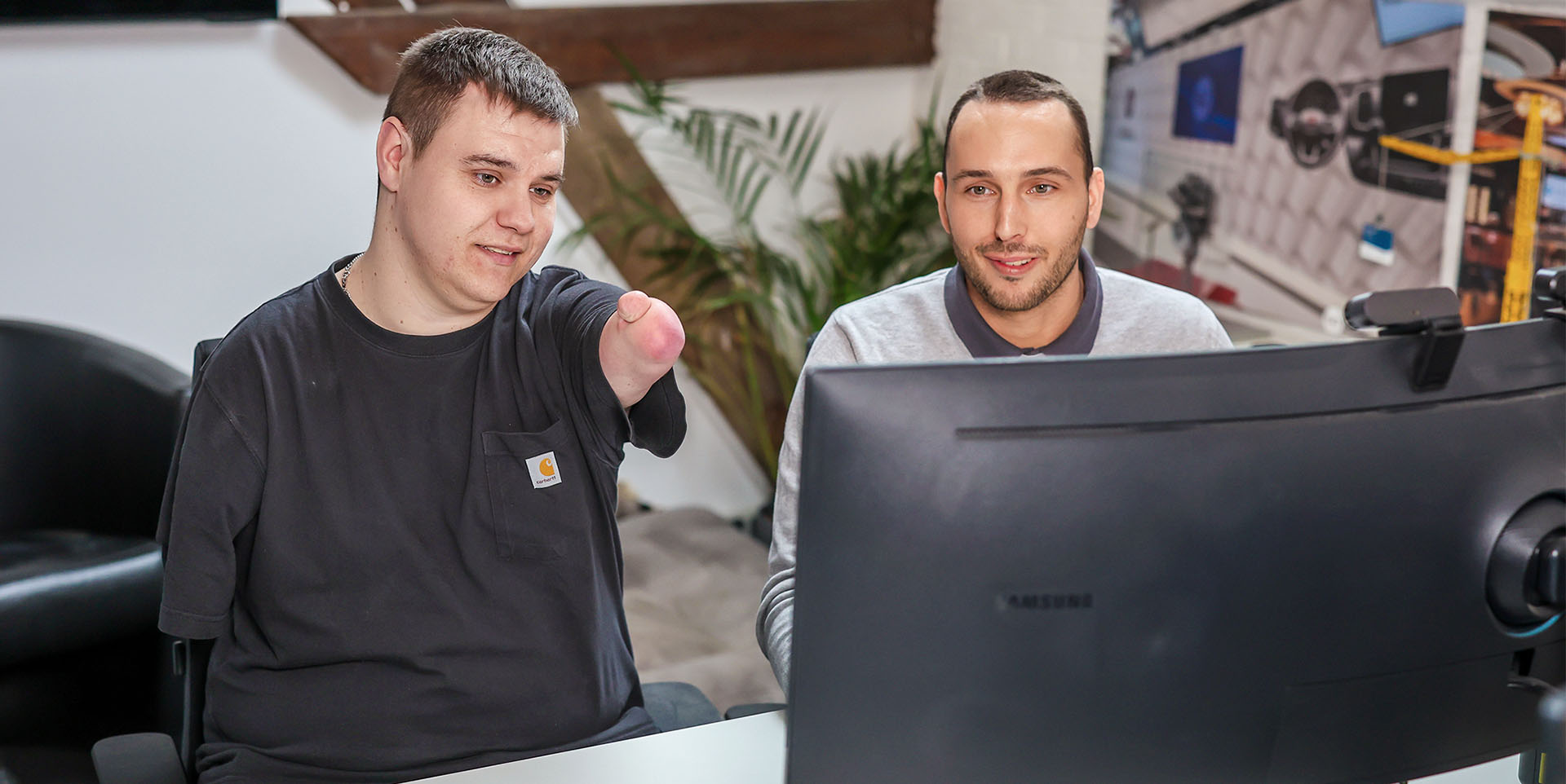 Yannik Boos (li.) und Benedikt Nöchelsitzen in einem modern eingerichteten Büro hinter einem großen Monitor. Yannik Boos zeigt mit seinem verkürzten Arm auf den Monitor. Beide lächeln.