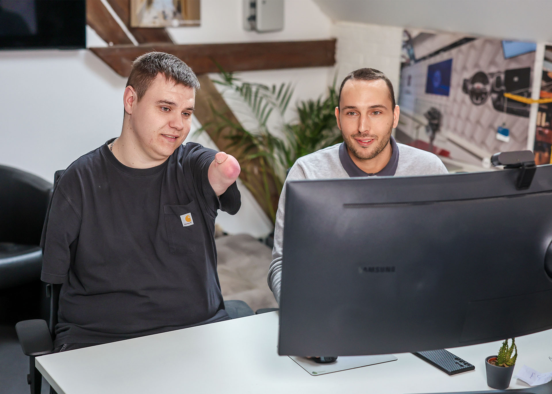 Yannik Boos (li.) und Benedikt Nöchelsitzen in einem modern eingerichteten Büro hinter einem großen Monitor. Yannik Boos zeigt mit seinem verkürzten Arm auf den Monitor. Beide lächeln.
