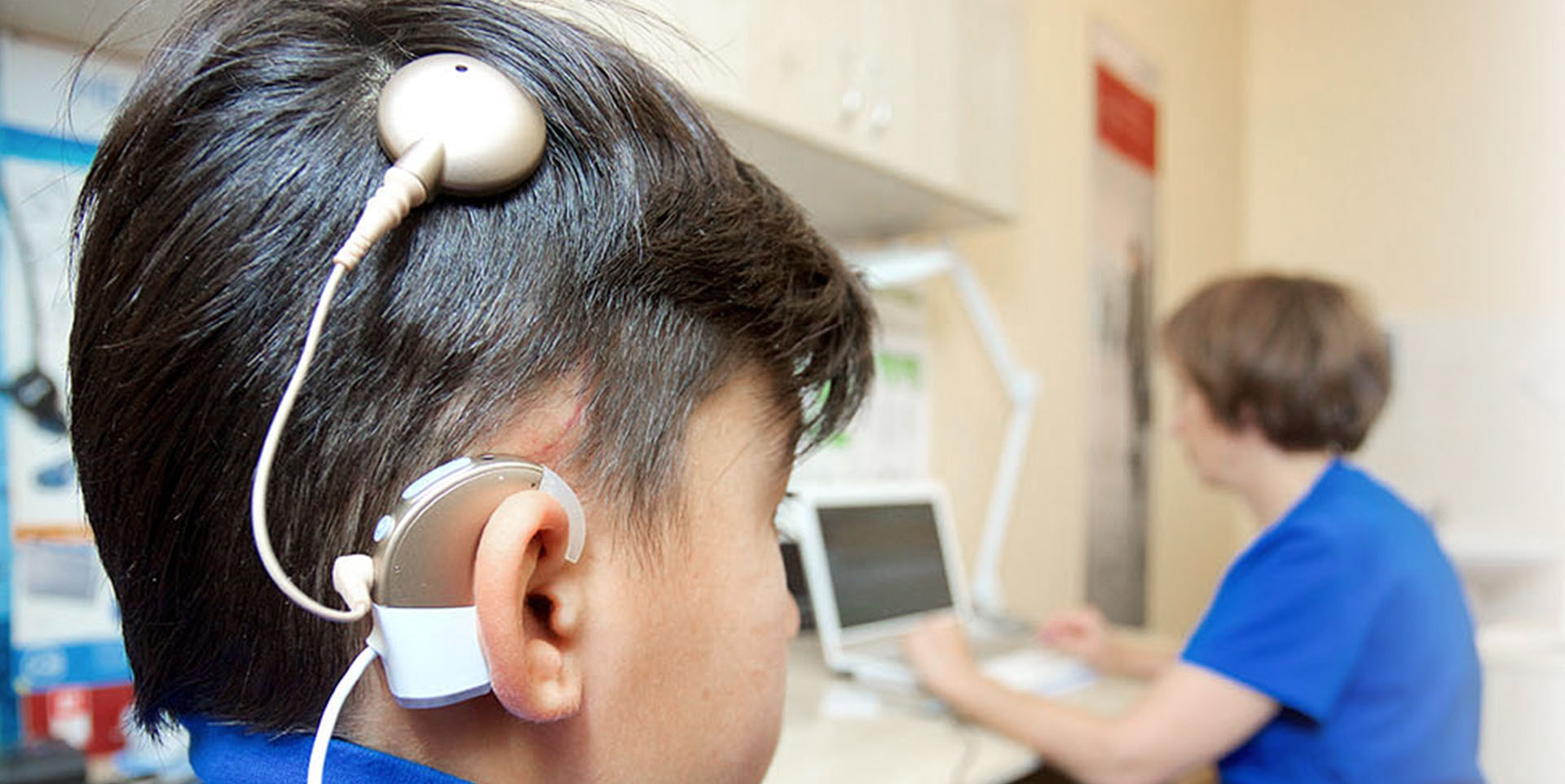 Cochlea-Implantat Ein Kind mit einem Cochlea-Implantat sitzt in einem medizinischen Untersuchungsraum, im Hintergrund ist eine Pflegekraft am Computer zu sehen.