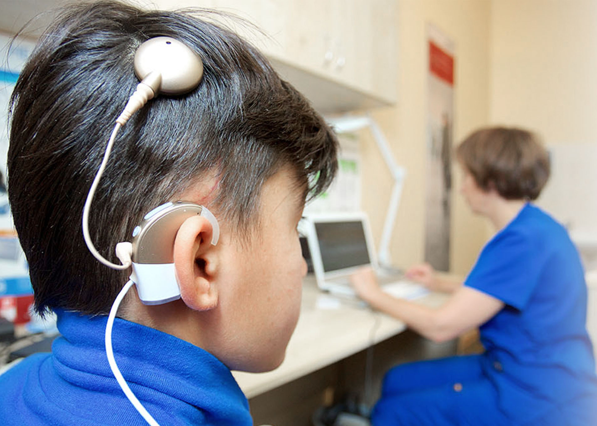 Ein Kind mit einem Cochlea-Implantat sitzt in einem medizinischen Untersuchungsraum, im Hintergrund ist eine Pflegekraft am Computer zu sehen.