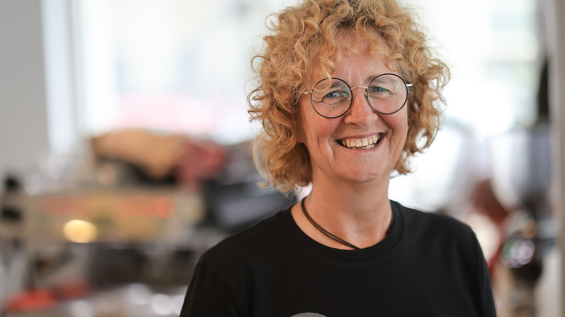 Eine Frau mittleren Alters mit rotblonden Locken und schwarzem T-Shirt lächelt direkt in die Kamera. Auf dem T-Shirt steht "signcafe. Inklusive Kaffeebude"