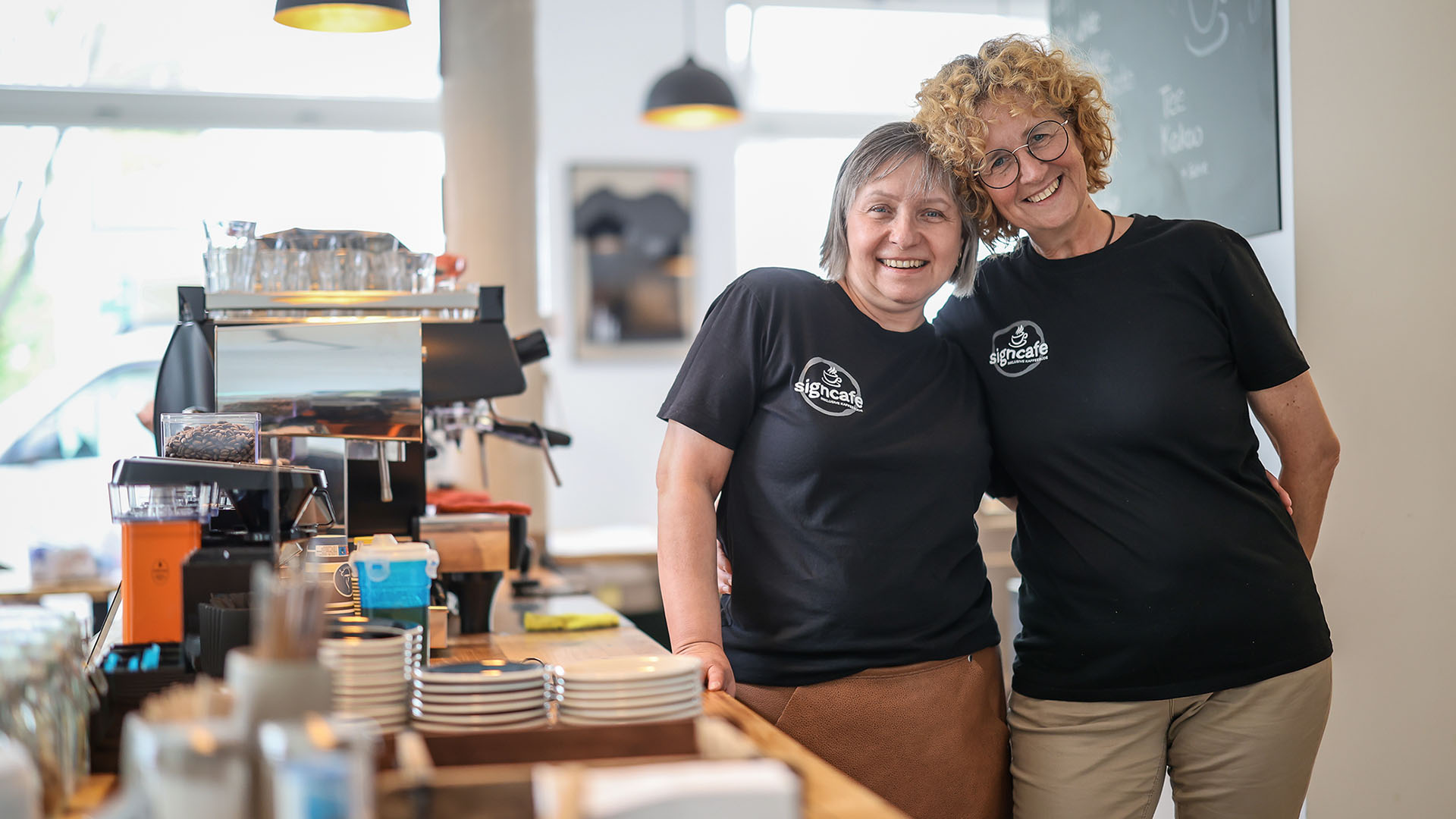 Zwei Frauen stehen Arm in Arm rechts von der Theke eines Cafés. Die Geschäftspartnerin legen lächelnd die Köpfe aneinander.