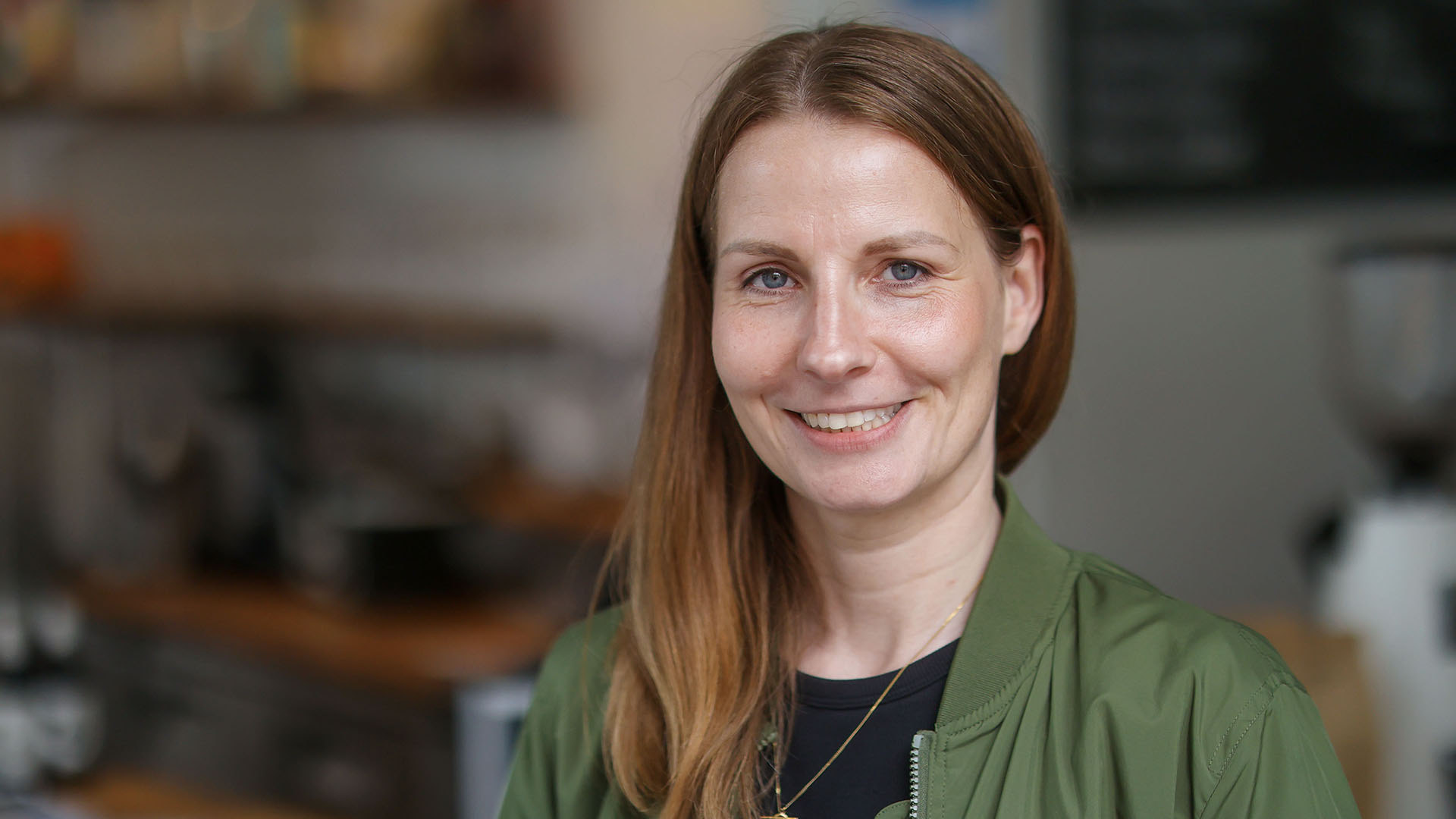 Eine junge Frau mit langen Haaren und grüner Blouson-Jacke steht lächelnd in einem Café.