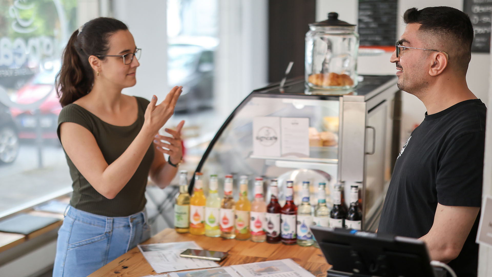 Eine Kuchentheke und Kasse in einem hellen, freundlichen Café. Eine junge Frau vor und ein junger Mann hinter der Theke unterhalten sich in deutscher Gebärdensprache. 