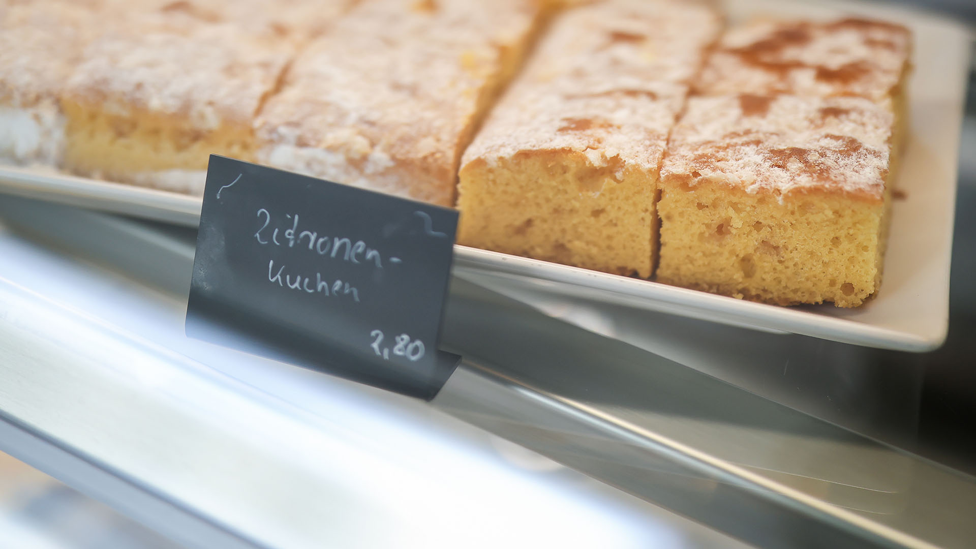 In der Kuchentheke eines Kaffees steht eine weiße längliche Platte mit Zitronenblechkuchen.