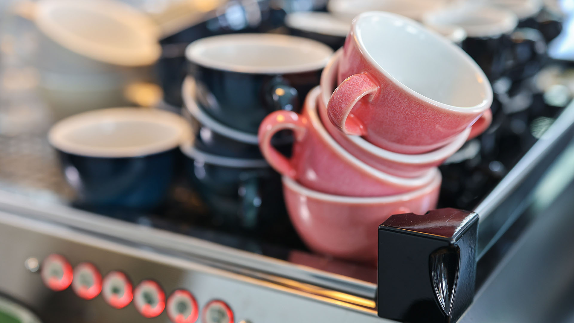 Blaue und lachsfarbene bauchige Kaffeetassen stehen in Stapeln geschützt von einer Reling auf einer großen Kaffeemaschine.