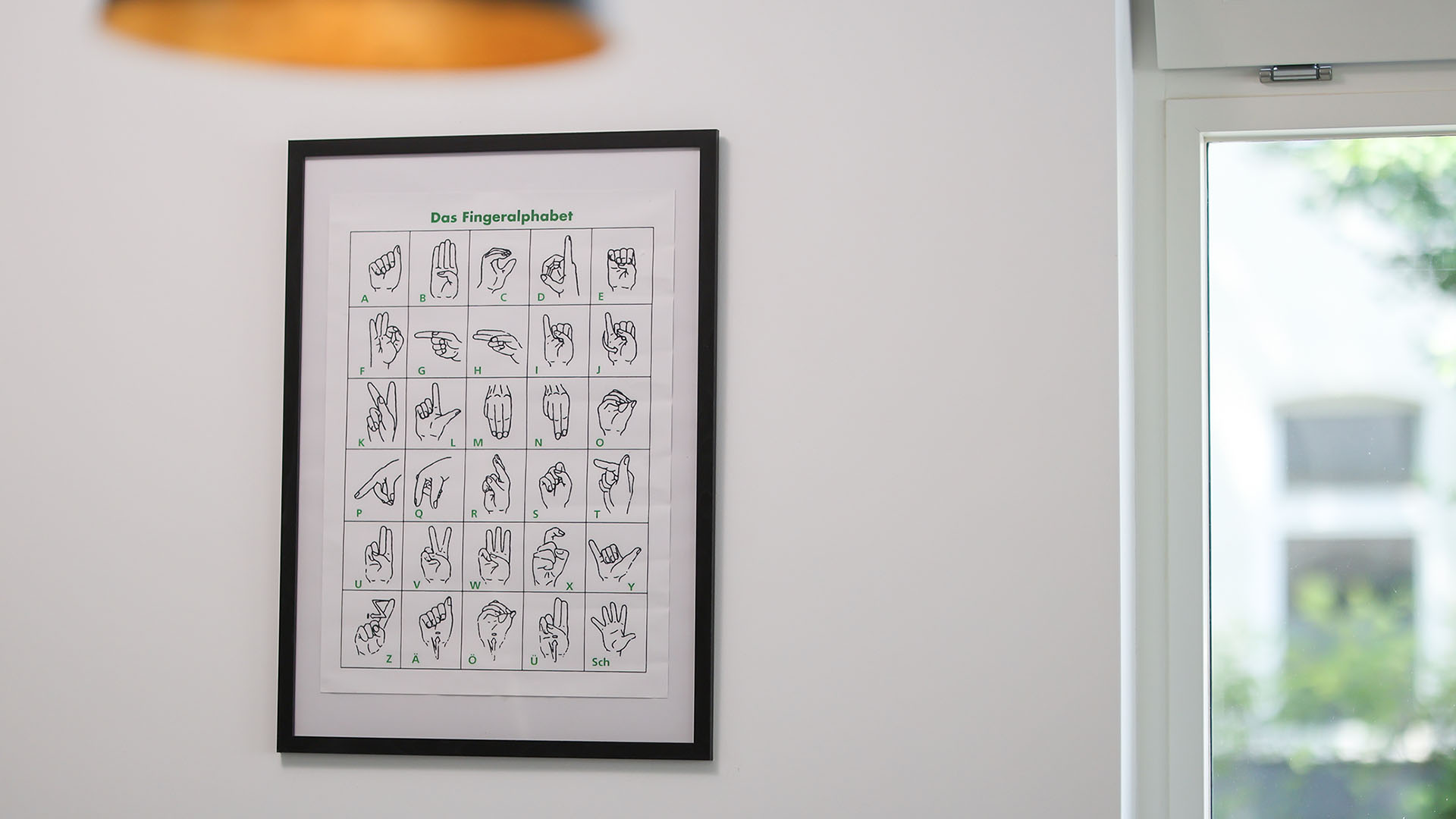 An einer weißen Wand hängt ein schwarzer Rahmen mit einem Poster. Darauf sind die Buchstaben der Gebärdensprache von A bis Z zu sehen.