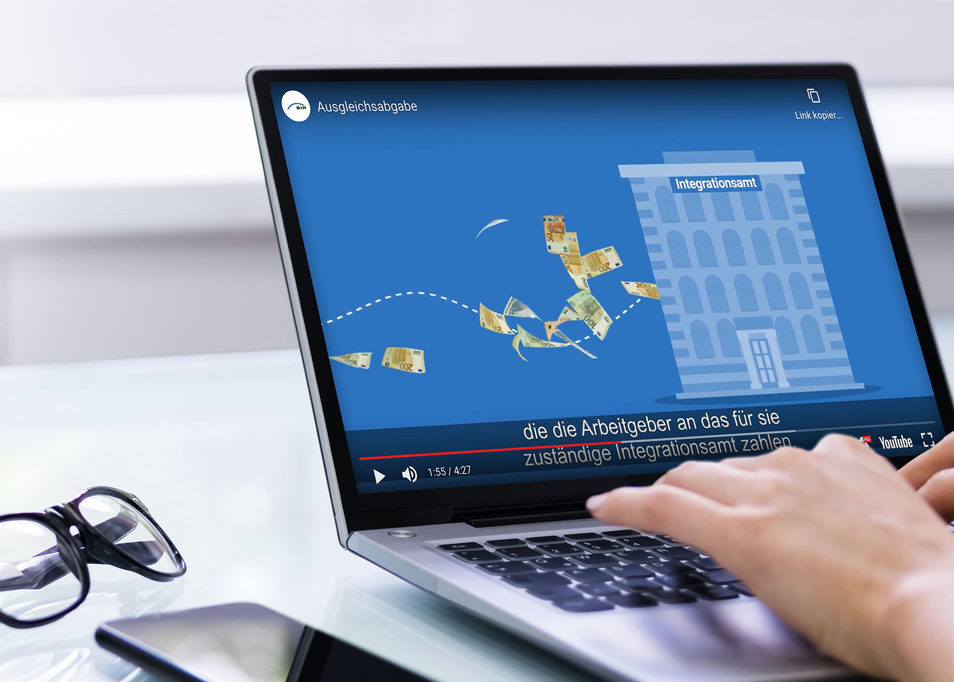 Auf einem Laptop-Monitor ist ein Ausschnitt eines Erklärfilms zur Ausgleichsabgabe zu sehen. In der Szene fliegen Geldscheine in ein Gebäude, das die Inklusions- und Integrationsämter symbolisiert.