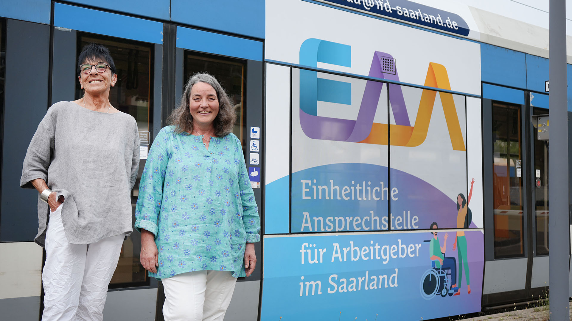 Kisten-Susanne-Dier (rechts) steht vor einem Zugwaggon, der mit dem Logo der EAA Saarland bedruckt ist.