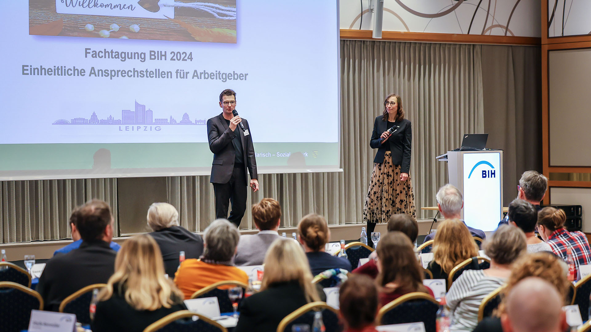 BIH-Vorstandsvorsitzender Christoph Beyer steht auf einer großen Bühne vor einem vollbesetzen Saal und spricht in ein Hand-Mikrophon. Im Hintergrund ist eine Präsentation an die Wand geworfen.