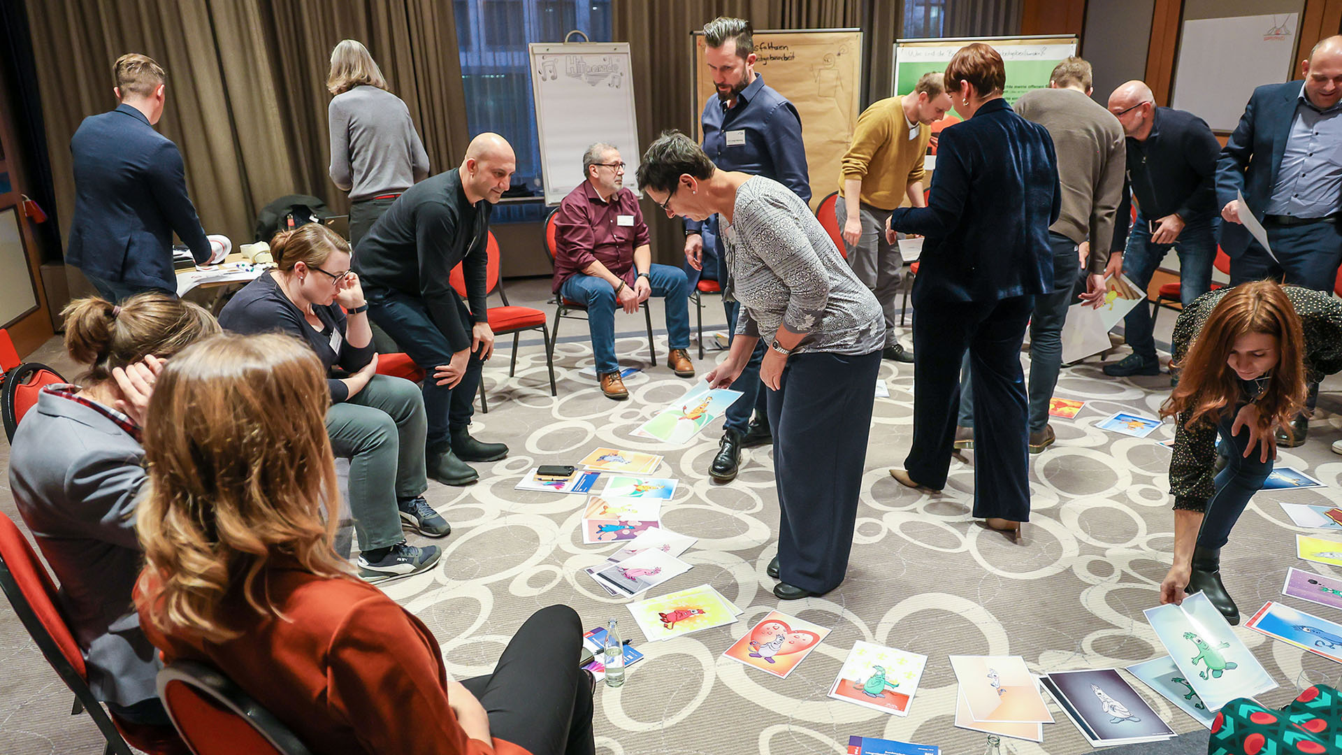 Teilnehmer des Workshops sitzen in einem Stuhlkreis. Mehrere Teilnehmer stehen in der Mitte des Kreises, während weitere Teilnehmer im Hintergrund die Notizen auf den dort aufgestellten Flip-Charts lesen.