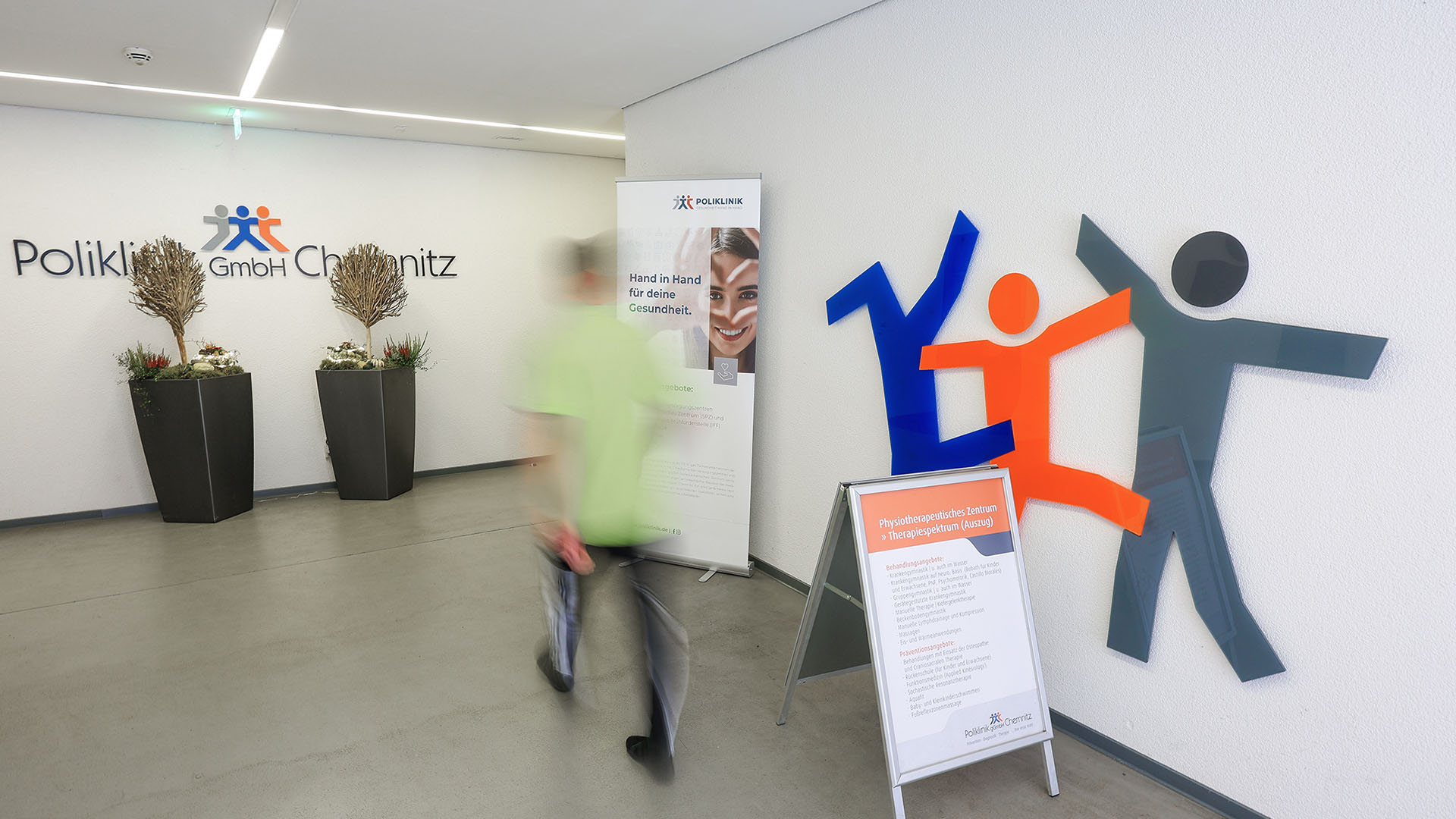 Ein junger Mann in Sportkleidung geht durch einen hellen, geräumigen Flur. An den Wänden sind bunte Zeichnungen von Strichmännchen in Bewegung angebracht. 