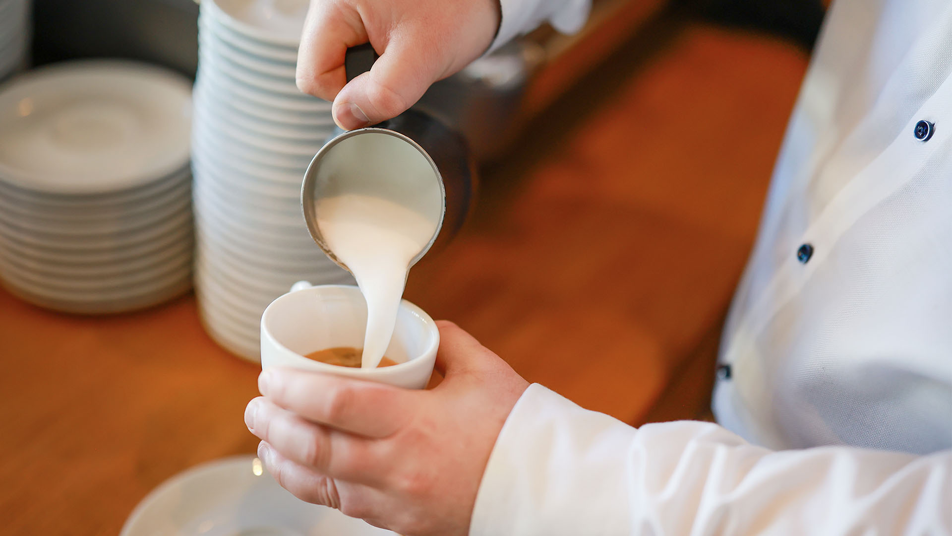 Nahaufnahme: aus einem kleinen Kännchen wird Milchschaum in eine Kaffeetasse gegossen.