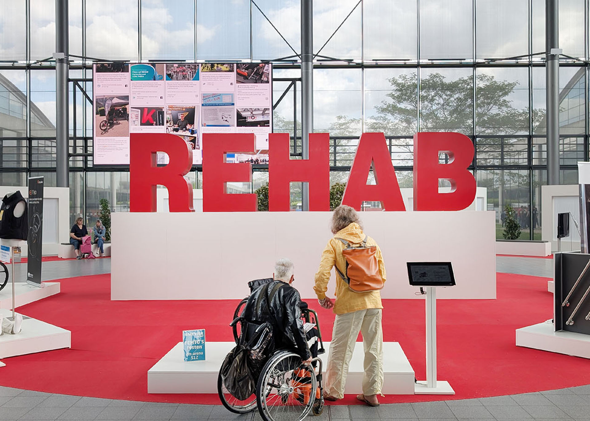 Großer Schriftzug der REHAB mit zwei Menschen davor, diese sieht man von hinten. 