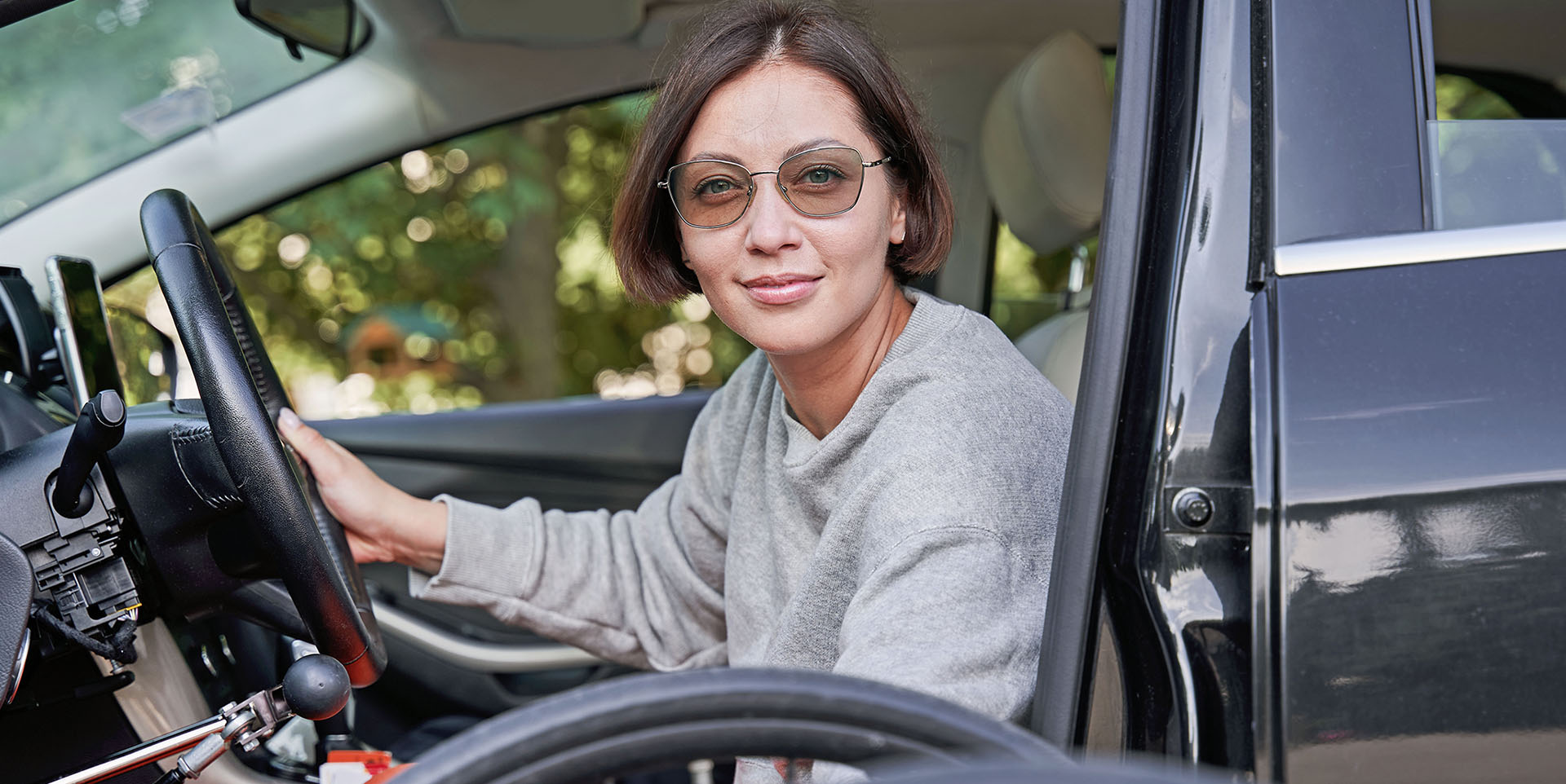Kfz-Umbau Eine junge Frau sitzt in einem umgebauten Kfz. Ihr Rollstuhl steht neben der geöffneten Fahrertür.