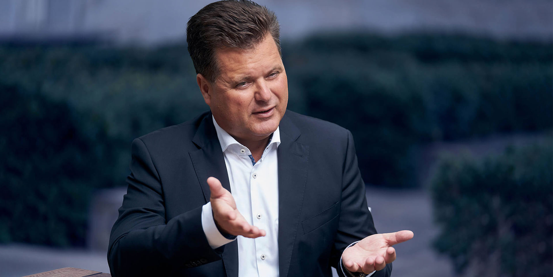 Jürgen Dusel sitzt an einem Tisch. Er trägt ein weißes Hemd und ein dunkelblaues Sakko. Er gestikuliert mit seinen Händen.