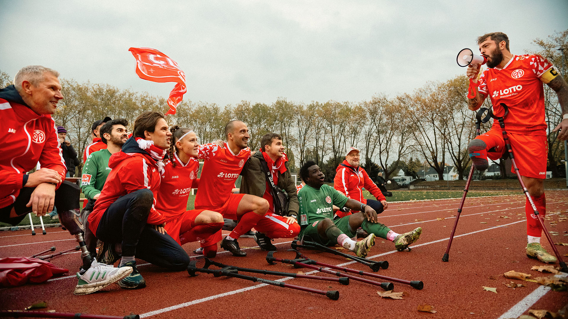 Ein Amputierten-Fußballmannschaft sitzt am Rand des Sportplatz, die Arme haben sie sich gegenseitig um die Schultern gelegt. Rechts steht ein Spieler mit einem Megaphone und hält eine Ansprache. 