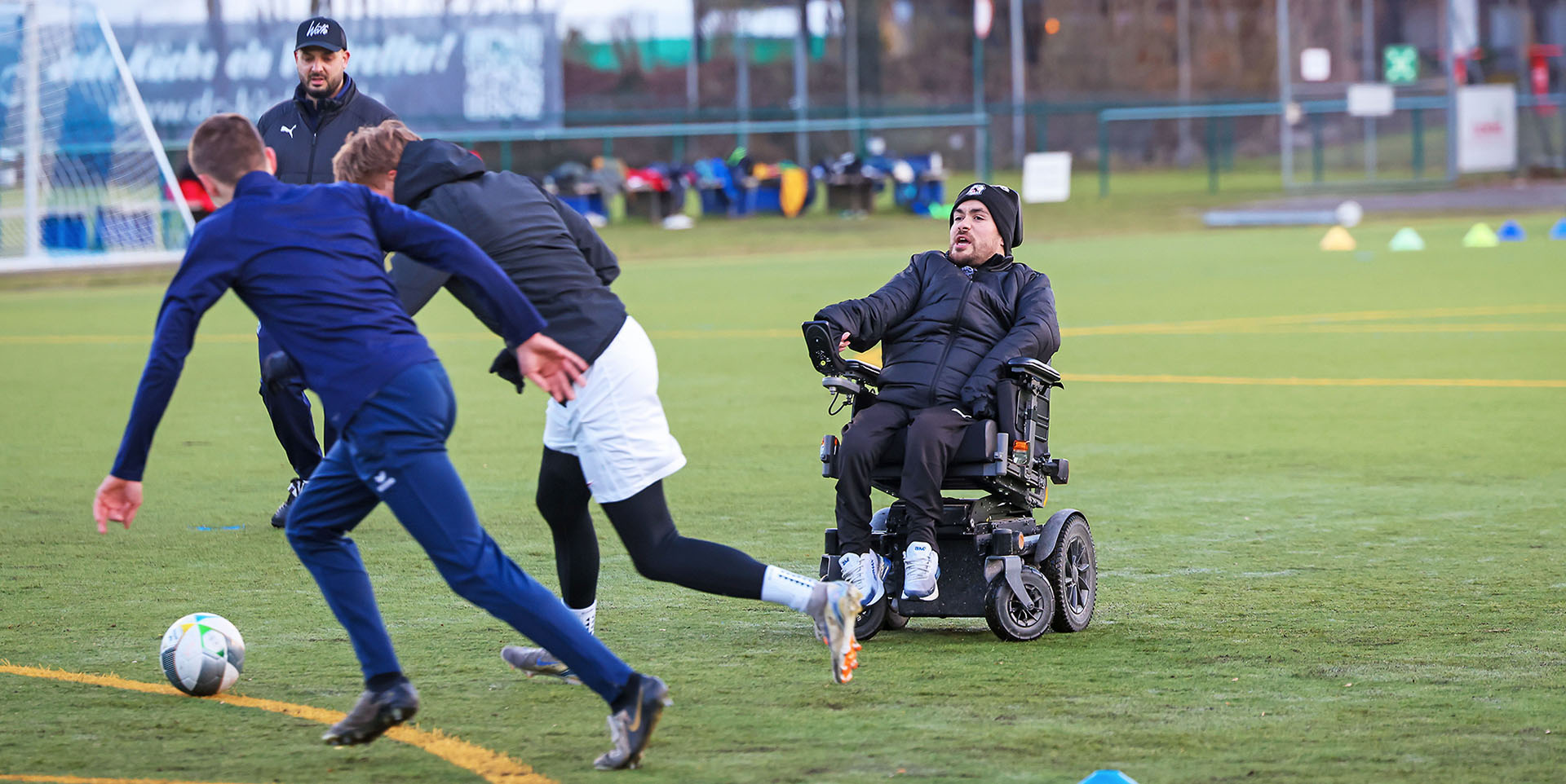 Behinderung im Sport Zwei Fußballer in Trainingsanzügen kämpfen um einen Ball. Dicht dahinter sind zwei Trainer – einer von beiden sitzt im Rollstuhl.