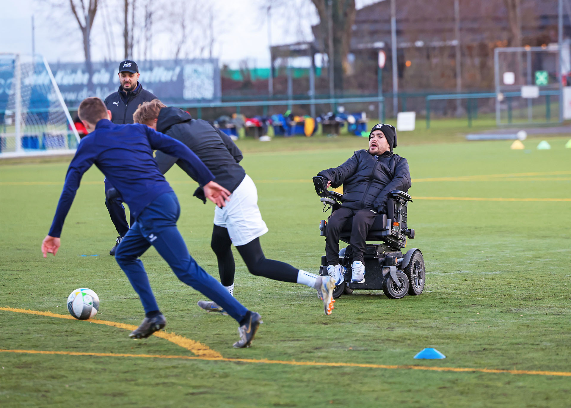 Zwei Fußballer in Trainingsanzügen kämpfen um einen Ball. Dicht dahinter sind zwei Trainer – einer von beiden sitzt im Rollstuhl.
