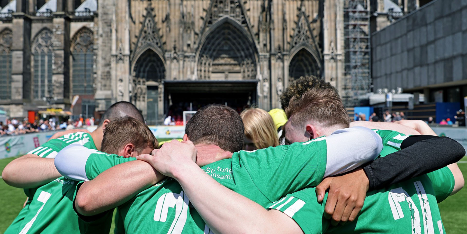 Eine Fußballmannschaft steht im Kreis und hat die Arme gegenseitig um die Schultern gelegt. Im Hintergrund ist der Kölner Dom zu sehen.
