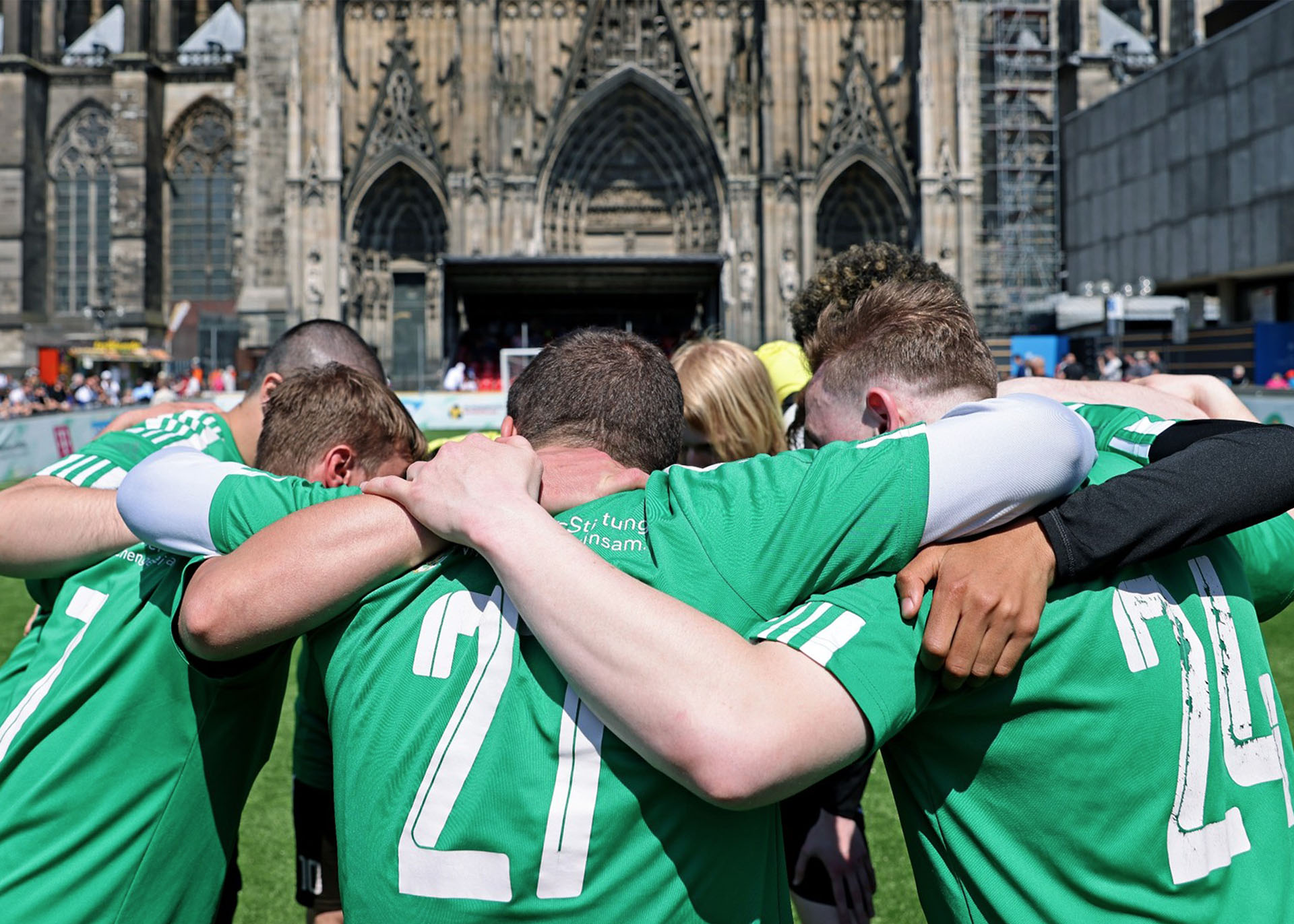 Eine Fußballmannschaft steht im Kreis und hat die Arme gegenseitig um die Schultern gelegt. Im Hintergrund ist der Kölner Dom zu sehen.