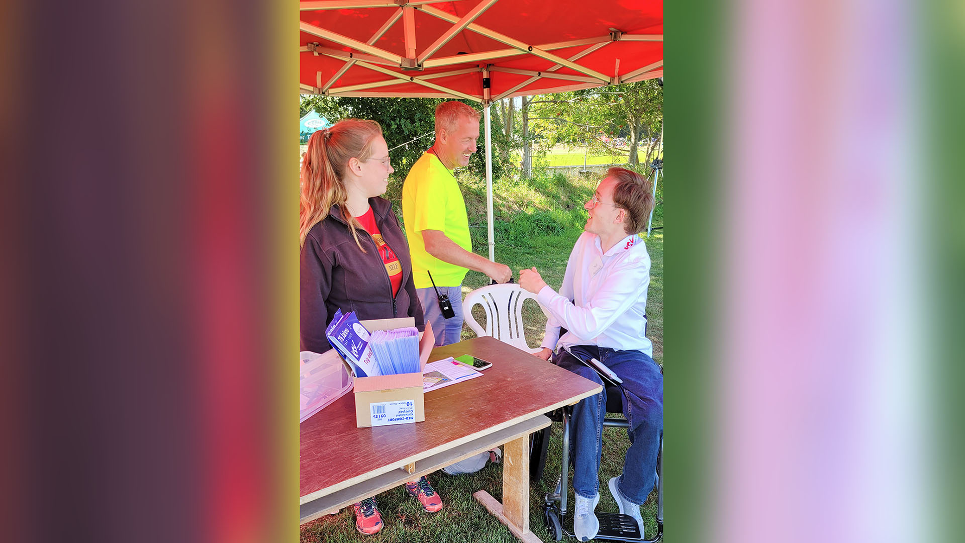 An einem Stand im Grünen unter einem Sonnenschirm begrüßt ein junger Mann im Rollstuhl einen anderen Mann, eine junge Frau steht daneben.  