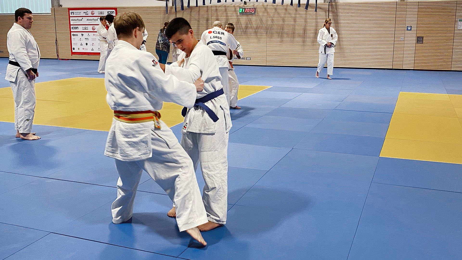 In einer Judo-Halle kämpfen zwei Jugendliche miteinander. Im Hintergrund sieht man weitere Judokas. 