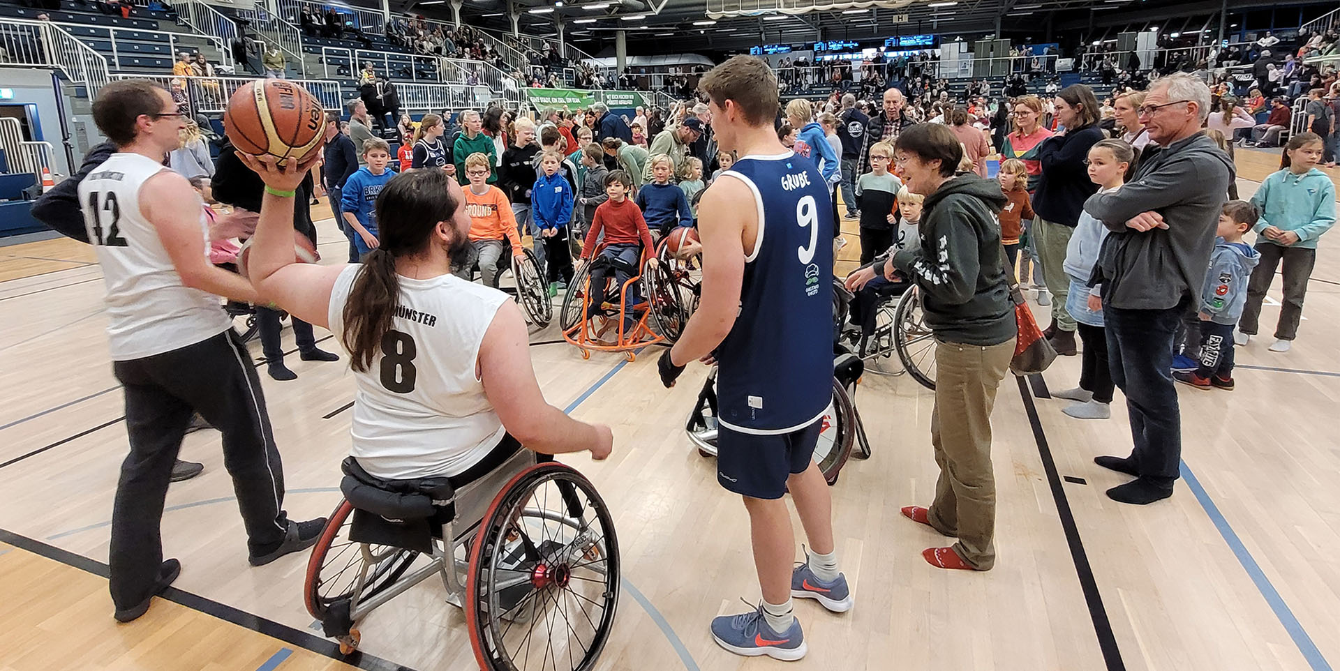 Auf dem Basketball-Feld stehen in viele Kinder im Rollstuhl einem Rollstuhl-Basketballer gegenüber, der einen Ball hochhält und ihnen etwas erklärt. 