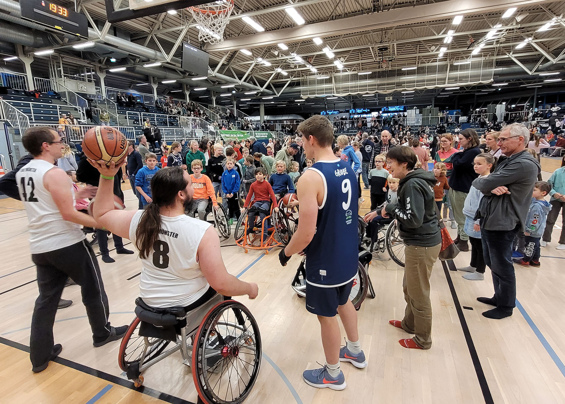 Auf dem Basketball-Feld stehen in viele Kinder im Rollstuhl einem Rollstuhl-Basketballer gegenüber, der einen Ball hochhält und ihnen etwas erklärt. 