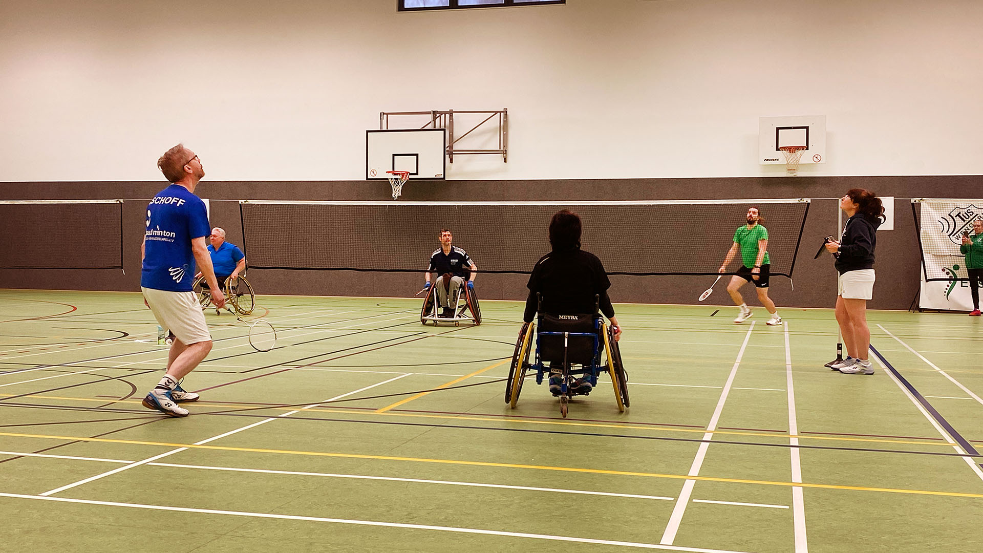 Ein Badmintonfeld: zwei gemischte Doppels - jeweils ein Spieler im Rollstuhl und ein Spieler ohne Gehbehinderung tragen ein Match aus.