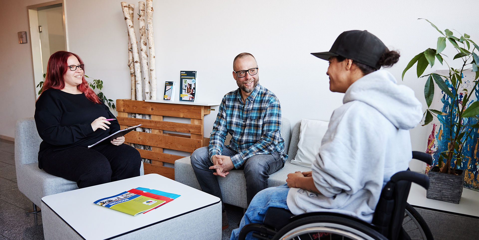Drei Menschen, eine Frau im Rollstuhl sitzen in einer Sitzgruppe bei einem Beratungsgespräch.