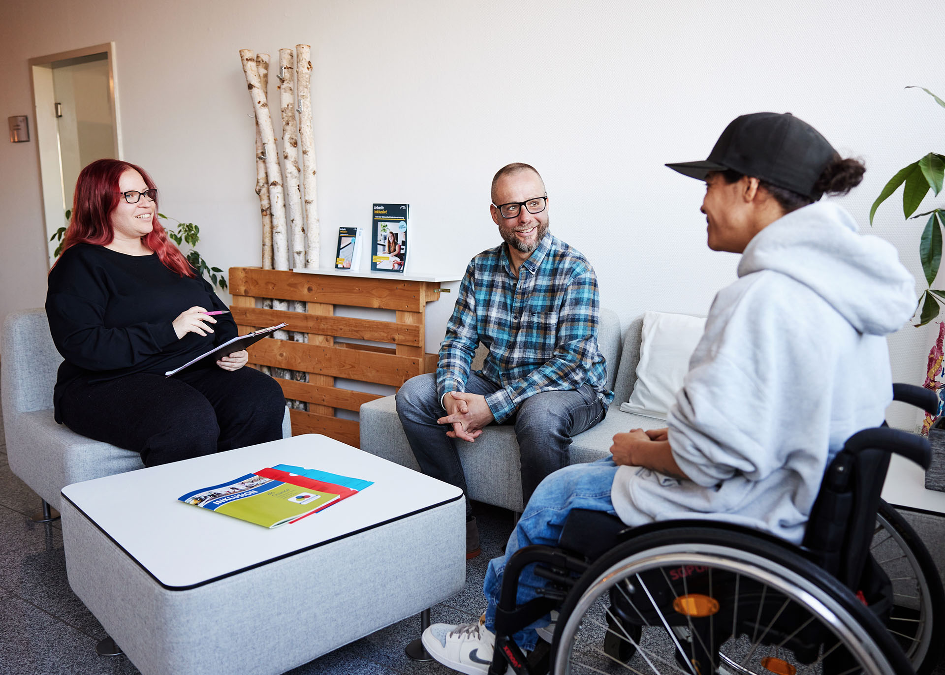 Drei Menschen, eine Frau im Rollstuhl sitzen in einer Sitzgruppe bei einem Beratungsgespräch. 