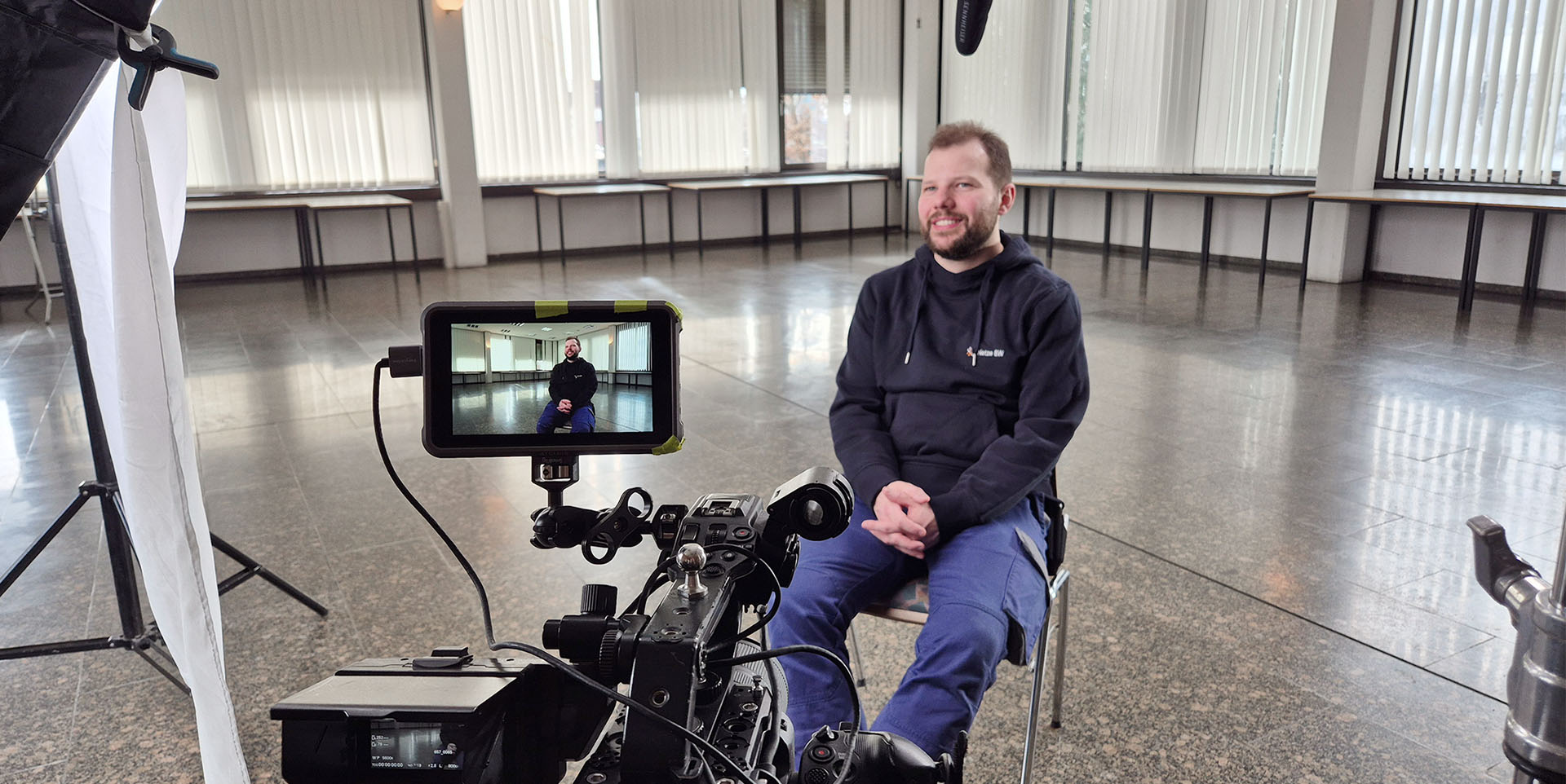 Ein Mann sitzt auf einem Stuhl, um ihn herum ist eine Interviewsituation für eine Filmaufnahme aufgebaut.