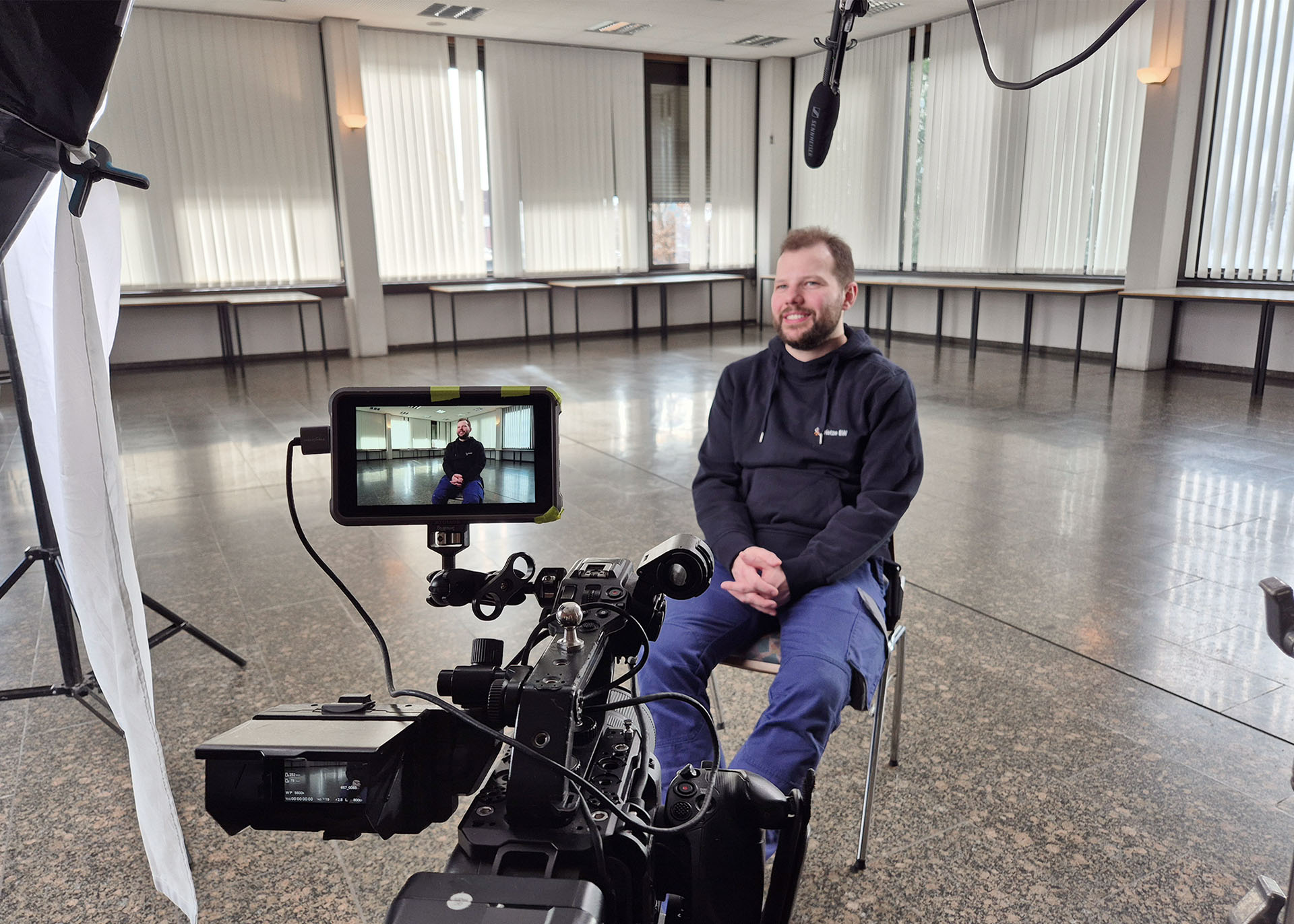 Ein Mann sitzt auf einem Stuhl, um ihn herum ist eine Interviewsituation für eine Filmaufnahme aufgebaut.
