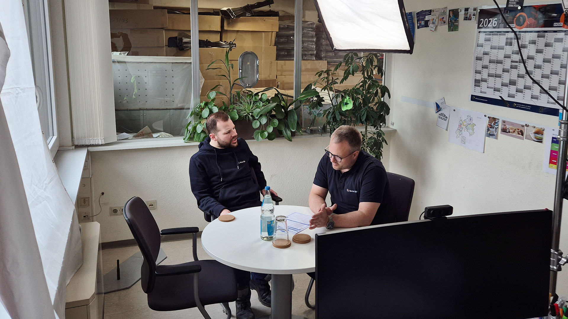 Zwei Männer sitzen an einem runden Besprechungstisch. Es sind Scheinwerfer und Kabel im Hintergrund zu sehen.