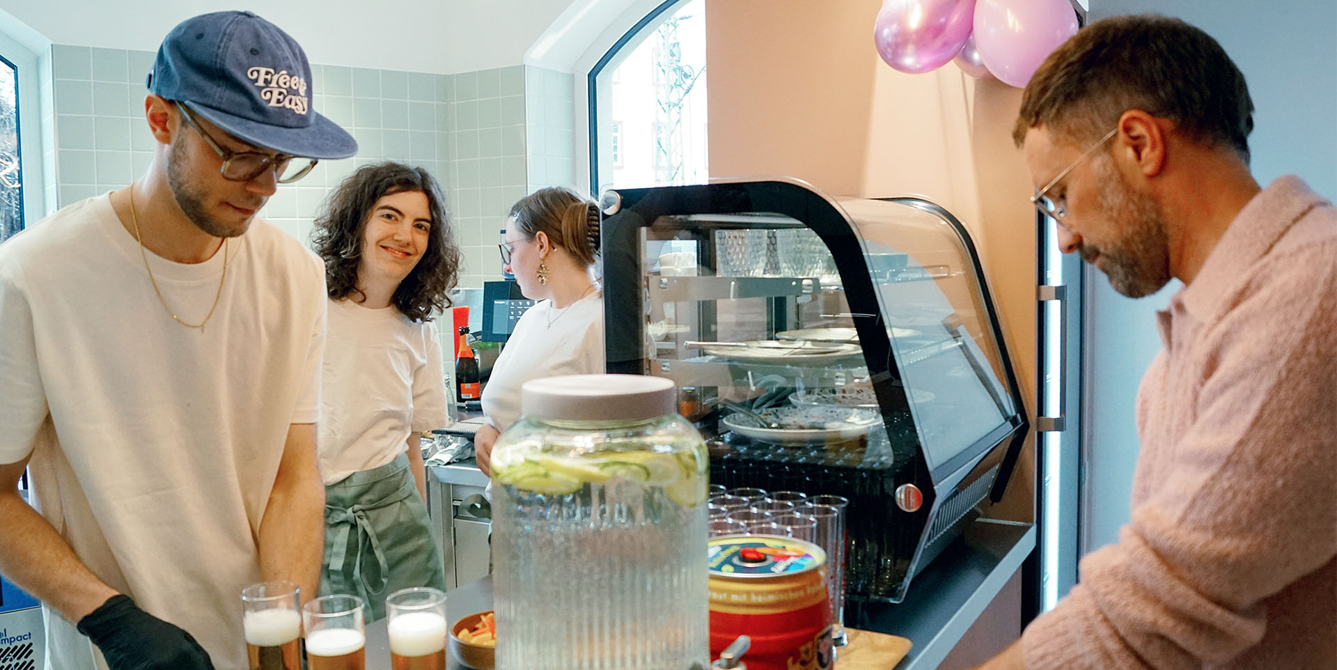 Eine Theke steht in einem hell gekacheltem und gestrichenen Café: drum herum vier junge Mitarbeitende – zwei Männer und zwei Frauen. 