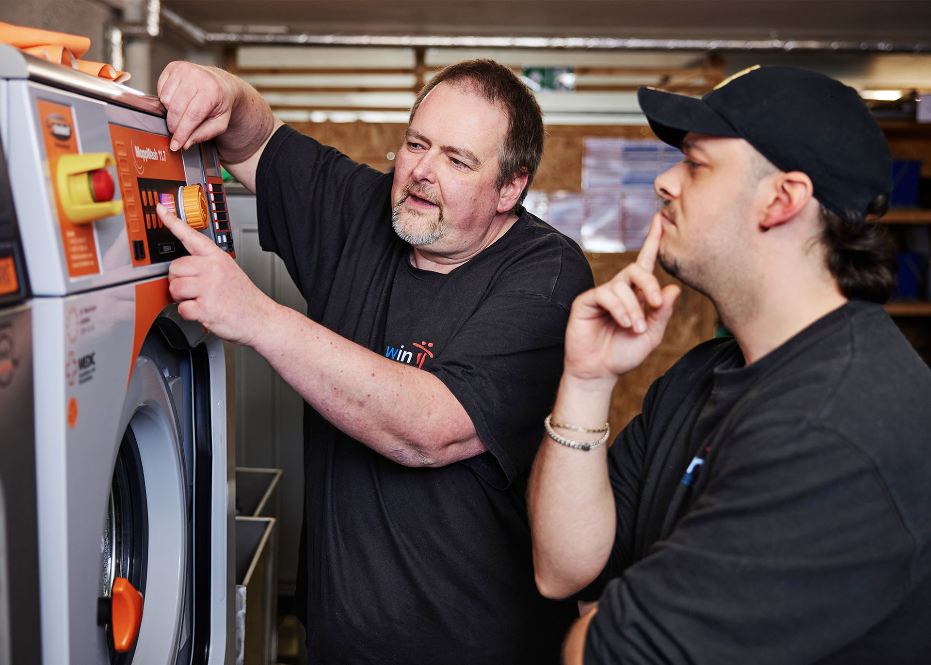 Zwei Männer stehen vor einer Industrie-Waschmaschine. Der ältere der beiden erklärt dem jüngeren die Bedienung.