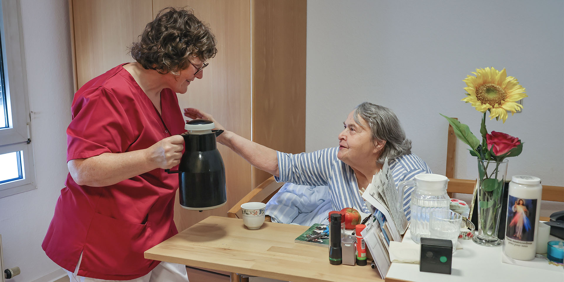 Jobcarving und Jobcoaching Eine weibliche Pflegekraft schenkt einer im Bett sitzenden Frau eine Tasse Kaffee ein.