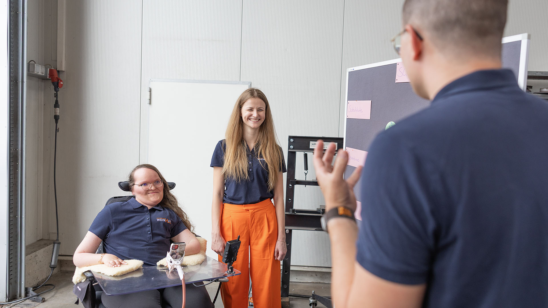 Patricia Lorenz, Julia Möslein und Andreas Weinhut stehen in einer Lagerhalle, in der sie Stellwände und Flippcharts für ihre Besprechung aufgebaut haben.