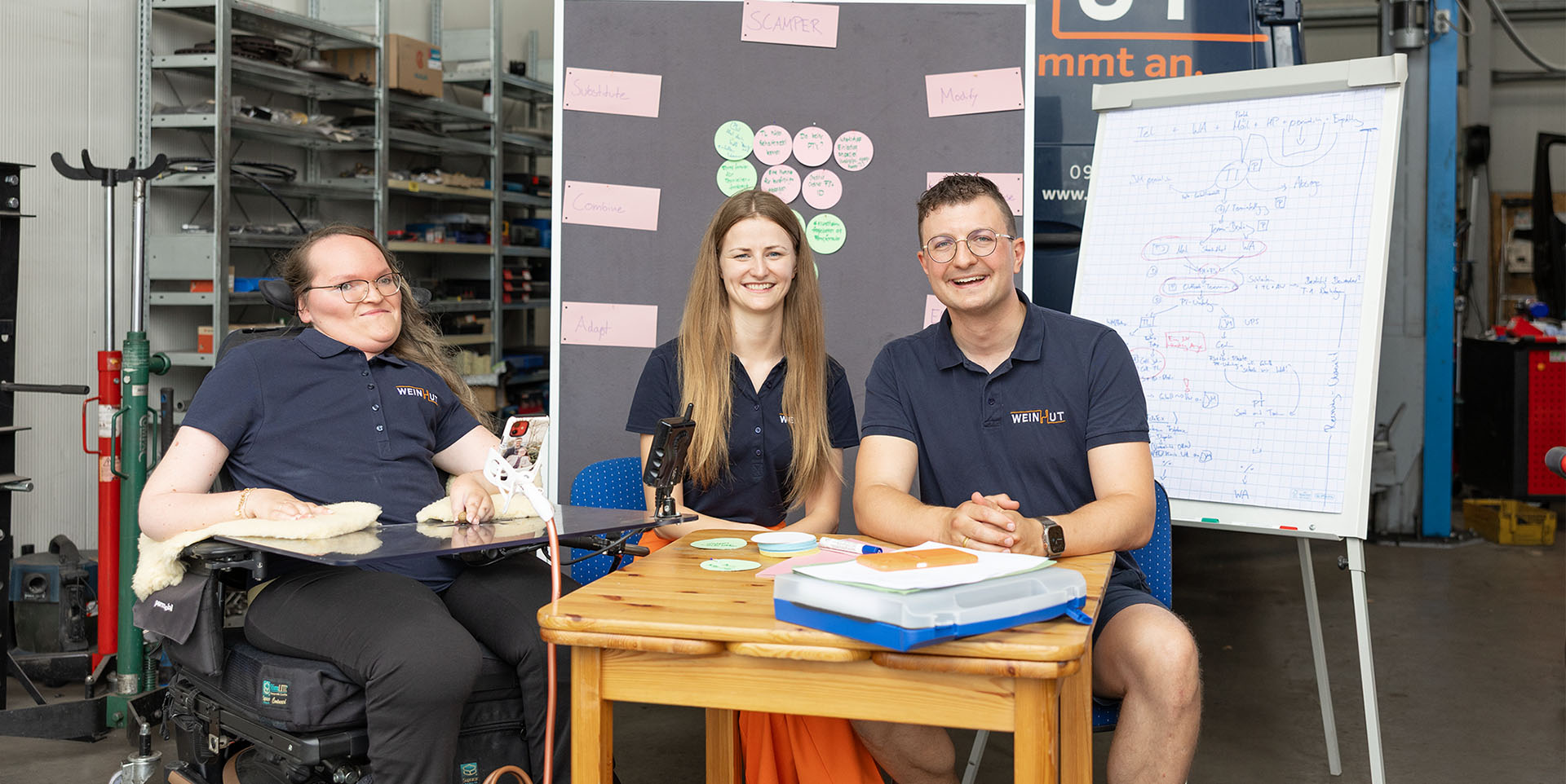 Patricia Lorenz, Julia Möslein und Andreas Weinhut sitzen an einem kleinen Tisch vor Flippcharts und Stellwänden. Im Hintergrund sind Lagerregale und Werkstattzubehör zu erkennen. 