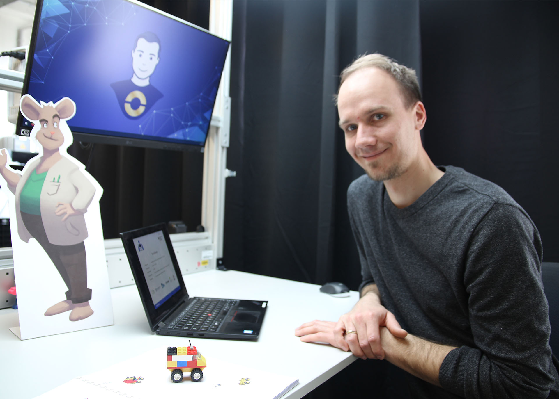 Der Projektkoordinator sitzt am Arbeitstisch. Darauf ein Laptop und ein Aufsteller der Comic-Maus Gregor im Anzug.